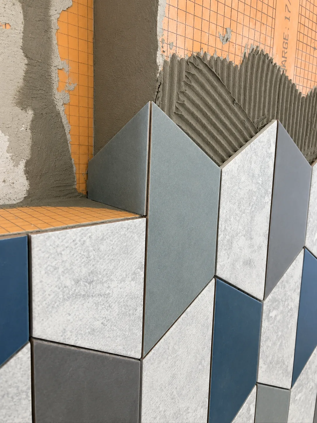 In-progress cube-pattern tile turning a mitred outside corner over orange waterproofing board, with combed thinset visible